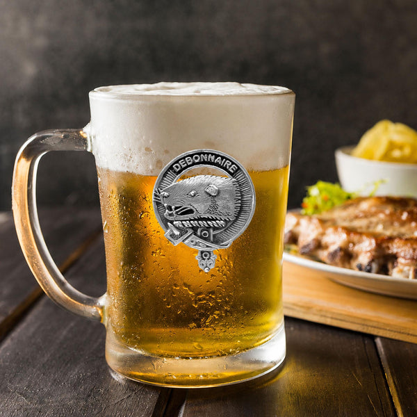 A glass mug filled with beer, featuring a silver emblem, sits on a wooden table next to a plate of food.