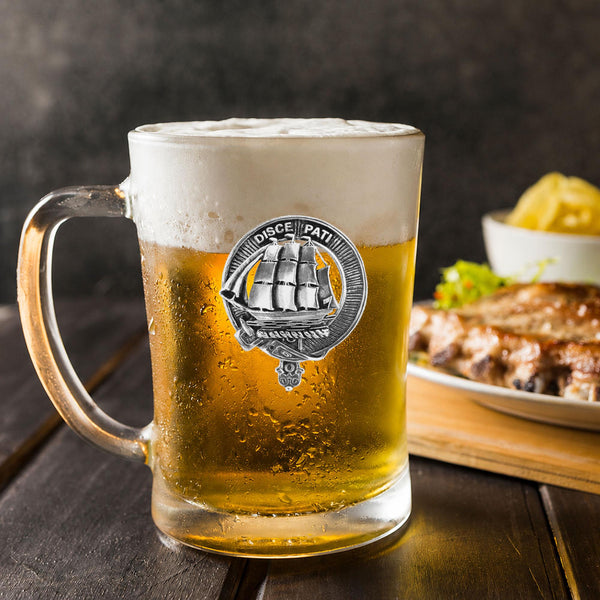 A glass mug filled with beer, featuring a silver ship emblem, sits on a wooden table next to a plate of food.