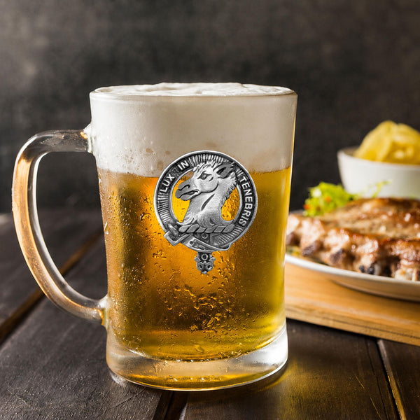 A glass mug filled with beer, featuring a silver emblem, sits on a wooden table next to a plate of food.