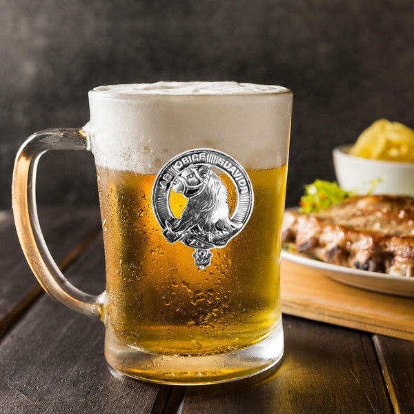 A glass mug filled with beer, featuring a silver emblem, sits on a wooden table next to a plate of food.