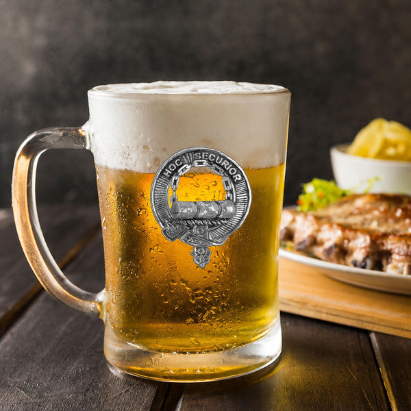 A glass mug filled with beer, featuring a silver emblem, sits on a wooden table next to a plate of food.