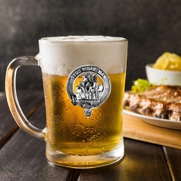 A glass mug filled with a golden-colored beer, topped with a frothy white head, sits on a wooden table. In the background, there is a plate of food, including what appears to be a piece of meat and some vegetables.