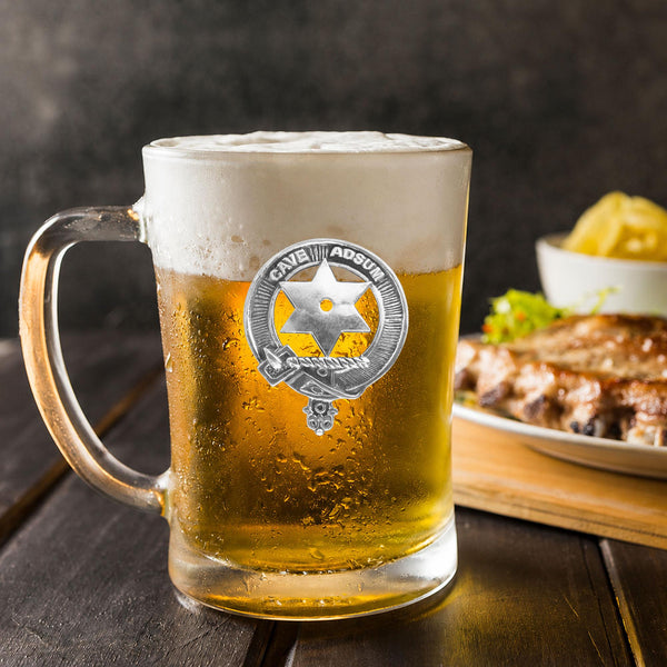 A glass mug filled with a golden-colored beer, topped with a frothy white head, sits on a wooden table. In the background, there is a plate of food, including what appears to be a piece of meat and some vegetables.