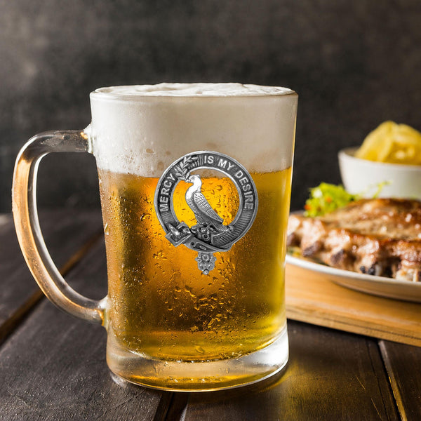 A glass mug filled with a golden-colored beer, topped with a frothy white head, sits on a wooden table. In the background, there is a plate of food, including what appears to be a piece of meat and some vegetables.