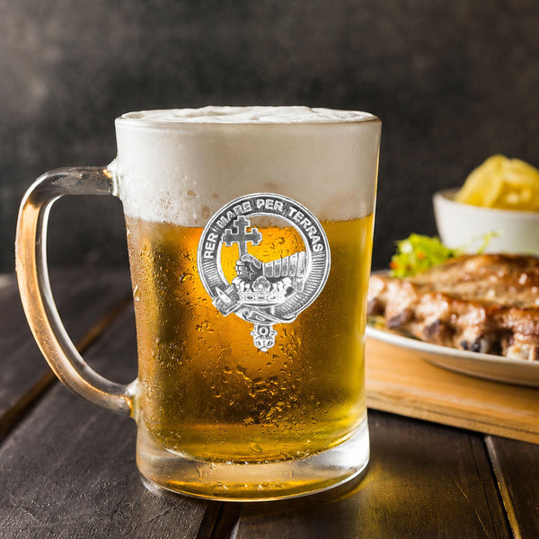 A glass mug filled with a golden-colored beer, topped with a frothy white head, sits on a wooden table. In the background, there is a plate of food, including what appears to be a piece of meat and some vegetables.