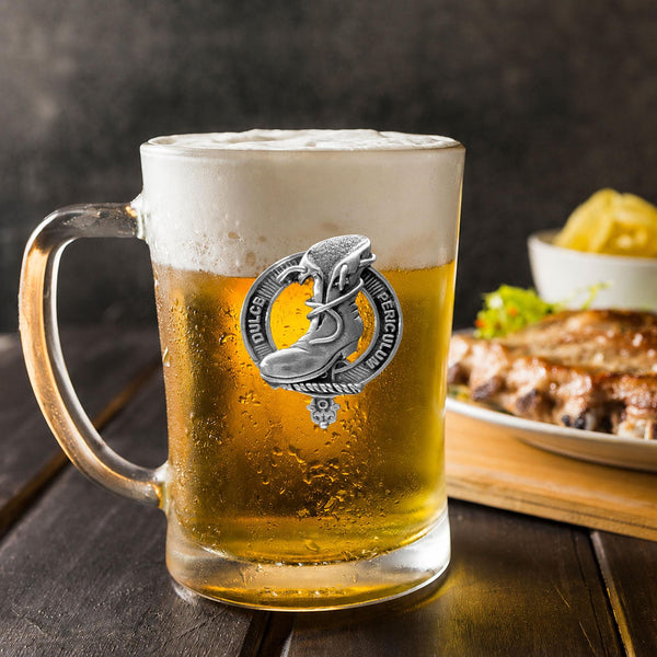 A glass mug filled with beer, featuring a silver emblem, sits on a wooden table next to a plate of food.