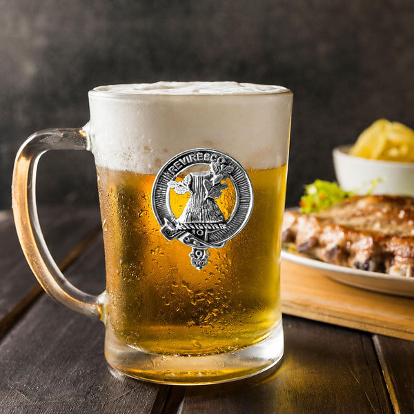 A glass mug filled with beer, featuring a metal emblem, sits on a wooden table next to a plate of food.