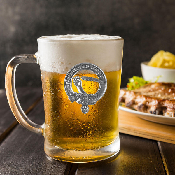 A glass mug filled with beer, featuring a silver emblem, sits on a wooden table next to a plate of food.