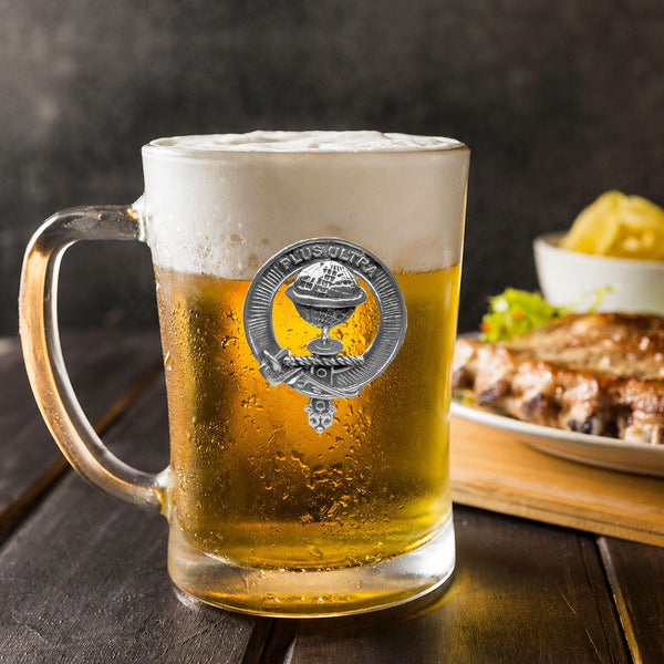 A glass mug filled with beer, featuring a silver emblem, sits on a wooden table next to a plate of food.