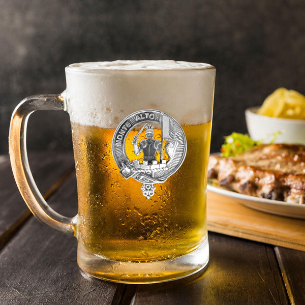 A glass mug filled with beer, featuring a silver badge with a coat of arms, placed on a wooden table. In the background, there is a plate of food, including what appears to be a piece of meat and some vegetables.