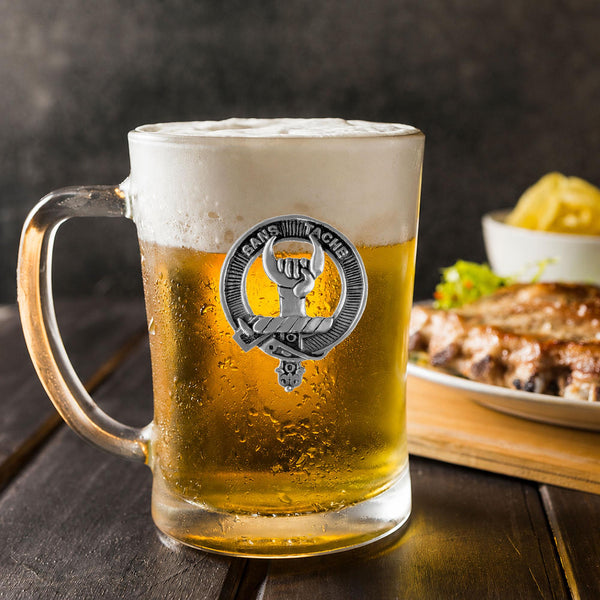 A glass mug filled with beer, featuring a silver emblem, sits on a wooden table next to a plate of food.