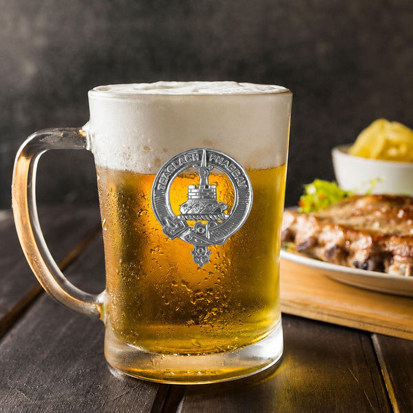 A glass mug filled with beer, featuring a silver emblem, sits on a wooden table next to a plate of food.