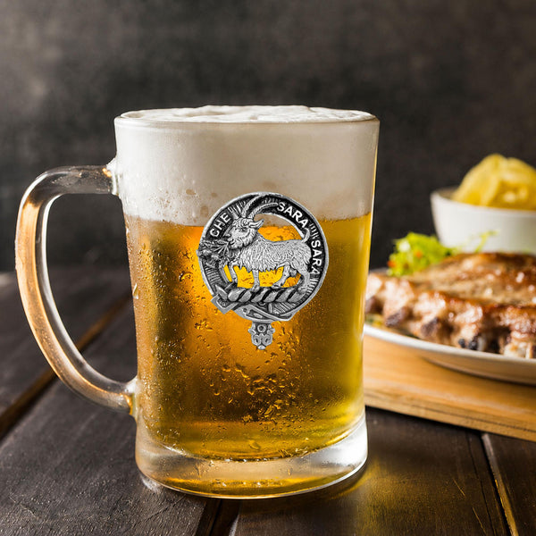 A glass mug filled with a golden-colored beer, featuring a logo, sits on a wooden table. In the background, there is a plate of food, including what appears to be a piece of meat and some vegetables.