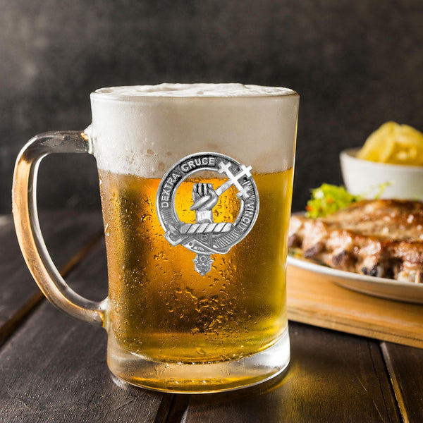 A glass mug filled with beer, featuring a silver emblem, sits on a wooden table next to a plate of food.