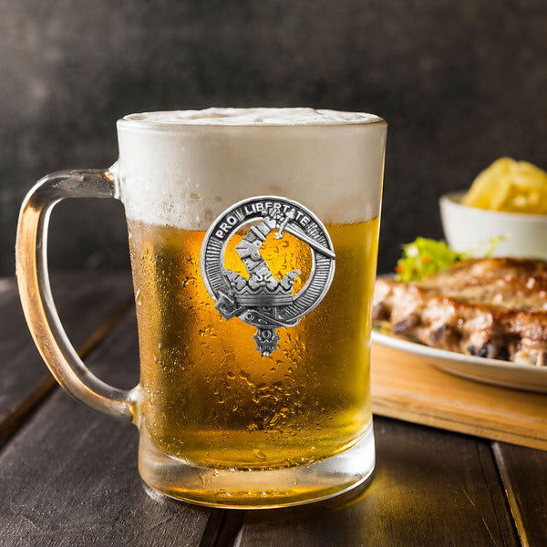 A glass mug filled with beer, featuring a silver emblem, sits on a wooden table next to a plate of food.