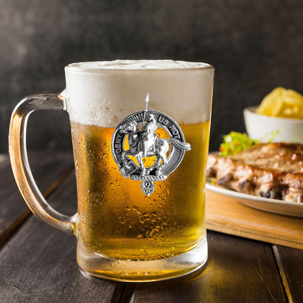 A glass mug filled with beer, featuring a silver emblem, sits on a wooden table next to a plate of food.