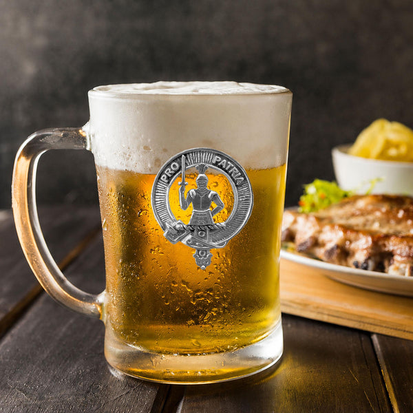 A glass mug filled with beer, featuring a silver badge with a figure and the text "PRO PATRIKA" on it, placed on a wooden table. In the background, there is a plate of food, including what appears to be a piece of meat and some vegetables.