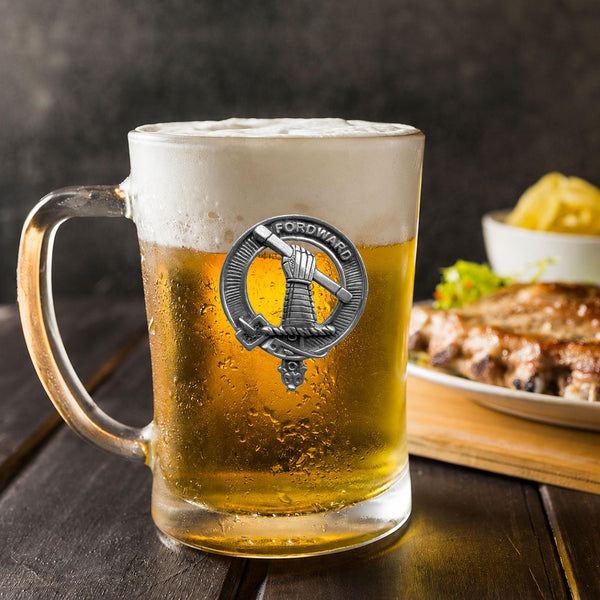 A glass mug filled with beer, featuring a metal emblem, sits on a wooden table next to a plate of food.