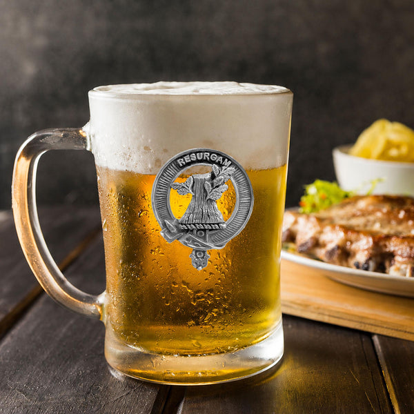 A glass mug filled with beer, featuring a silver emblem, sits on a wooden table next to a plate of food.