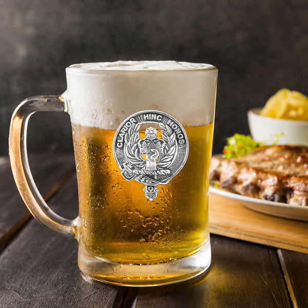 A glass mug filled with a golden-colored beer, topped with a frothy white head, sits on a wooden table. In the background, there is a plate of food, including what appears to be a piece of meat and some vegetables.