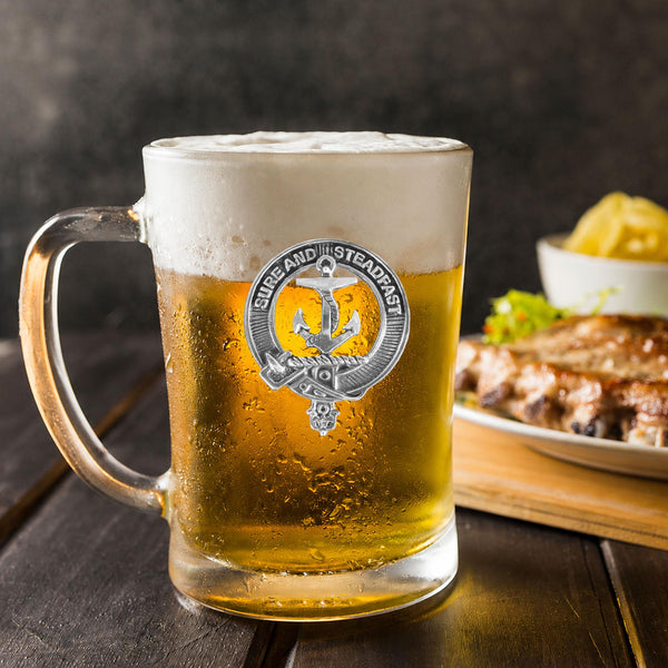 A glass mug filled with a golden-colored beer, topped with a frothy white head, sits on a wooden table. In the background, there is a plate of food, including what appears to be a piece of meat and some vegetables.