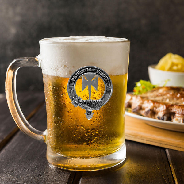 A glass mug filled with beer, featuring a silver badge with a logo, sits on a wooden table next to a plate of food.