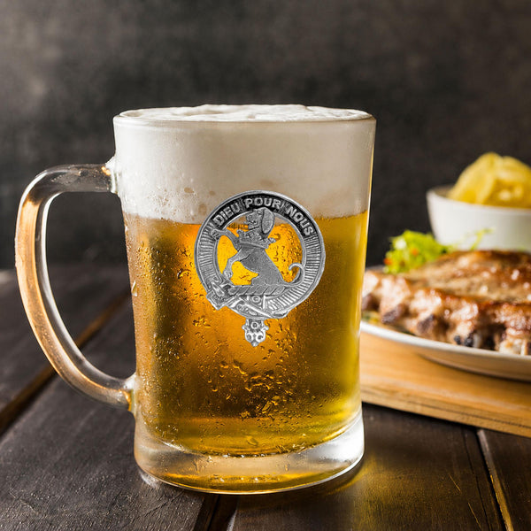 A glass mug filled with a golden-colored beer, topped with a frothy white head, sits on a wooden table. In the background, there is a plate of food, including what appears to be a piece of meat and some vegetables.