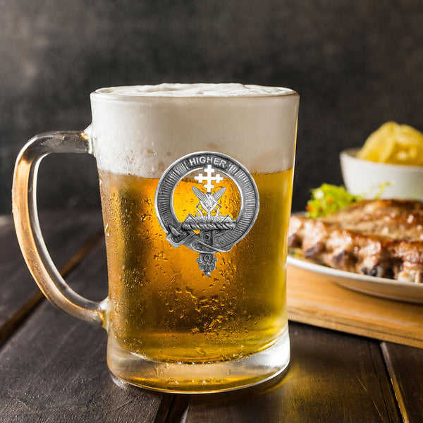 A glass mug filled with beer, featuring a silver badge with a crest and the text "HIGHER" on it, placed on a wooden table. In the background, there is a plate of food, including what appears to be a piece of meat and some vegetables.