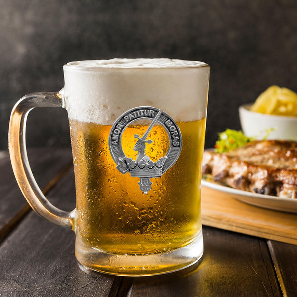 A glass mug filled with beer, featuring a silver emblem, sits on a wooden table next to a plate of food.