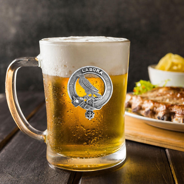 A glass mug filled with beer, featuring a metal emblem, sits on a wooden table next to a plate of food.