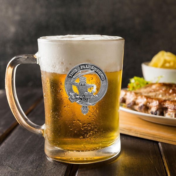 A glass mug filled with beer, featuring a silver emblem, sits on a wooden table next to a plate of food.
