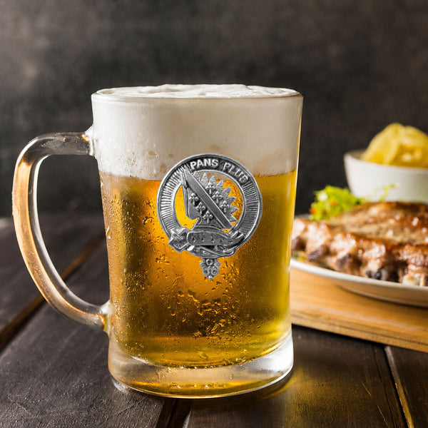 A glass mug filled with beer, featuring a silver emblem, sits on a wooden table next to a plate of food.