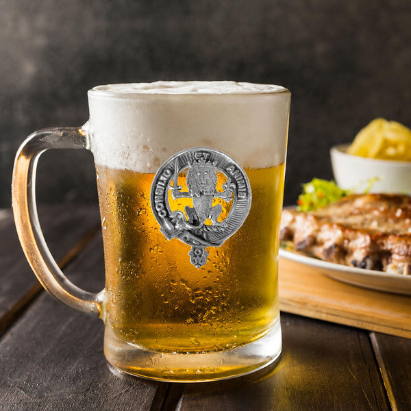 A glass mug filled with beer, featuring a silver emblem, sits on a wooden table next to a plate of food.
