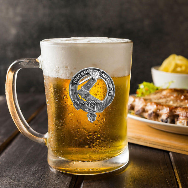 A glass mug filled with beer, featuring a silver emblem, sits on a wooden table next to a plate of food.