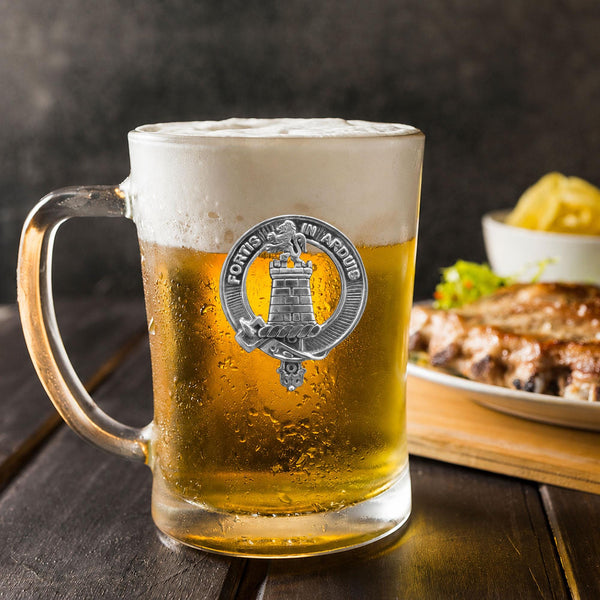A glass mug filled with beer, featuring a silver emblem, sits on a wooden table next to a plate of food.