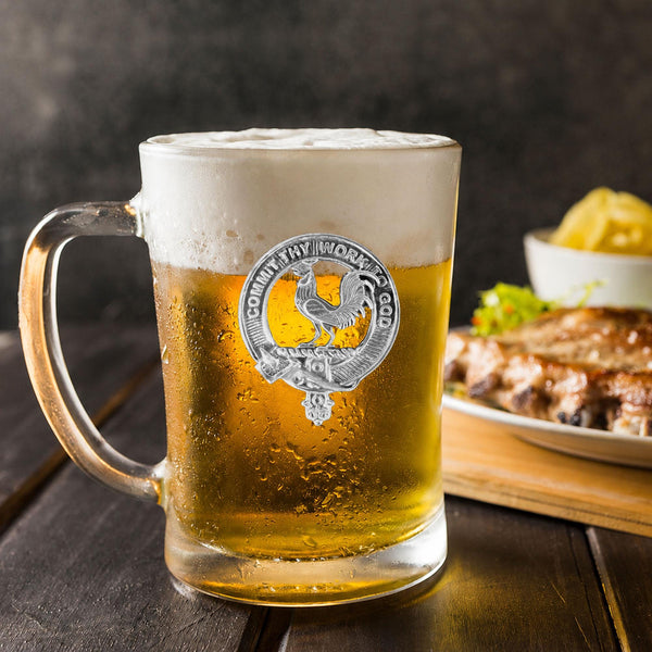 A glass mug filled with a golden-colored beer, topped with a frothy white head, sits on a wooden table. In the background, there is a plate of food, including what appears to be a piece of meat and some vegetables.