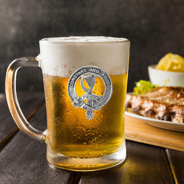 A glass mug filled with a golden-colored beer, topped with a frothy white head, sits on a wooden table. In the background, there is a plate of food, including what appears to be a piece of meat and some vegetables.
