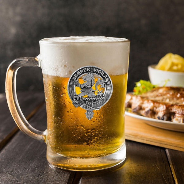 A glass mug filled with beer, featuring a silver emblem, sits on a wooden table next to a plate of food.