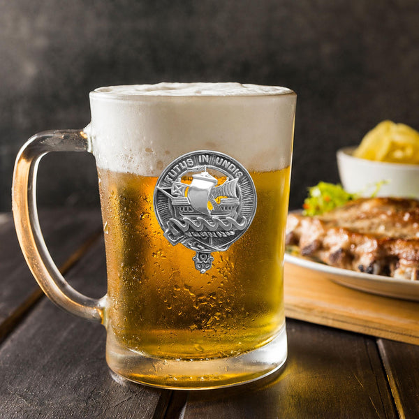 A glass mug filled with beer, featuring a silver emblem, sits on a wooden table next to a plate of food.